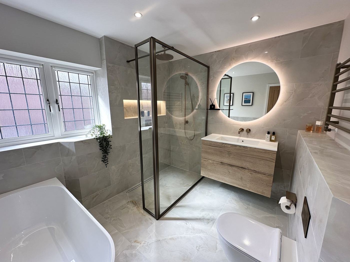 Bathroom renovation with a bronze-framed walk-in shower, backlit round mirror and floating oak vanity in Kleinburg, by NewGen Renovations Vaughan