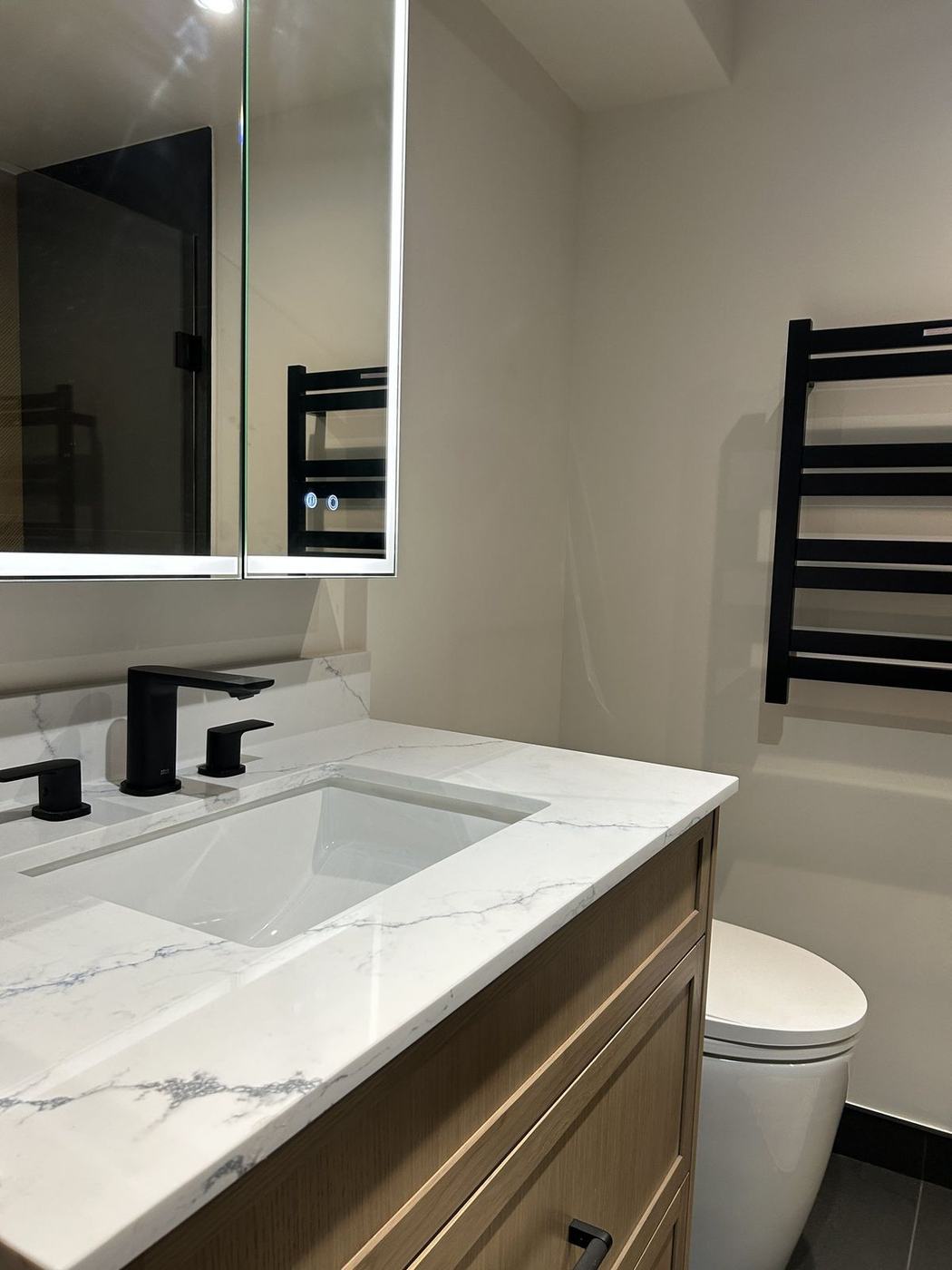 Renovated bathroom with an oak vanity, veined quartz countertop, matte black faucet, lit mirror cabinet and heated towel rack, by NewGen Renovations Vaughan